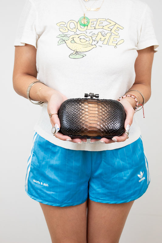 Person holding a black snake-textured clutch bag wearing a white t-shirt with a graphic and blue shorts.
