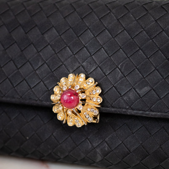 Black woven clutch with a decorative brooch featuring a pink gemstone and gold accents on a light background.