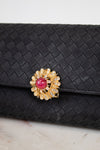 Black woven clutch with a decorative brooch featuring a pink gemstone and gold accents on a light background.