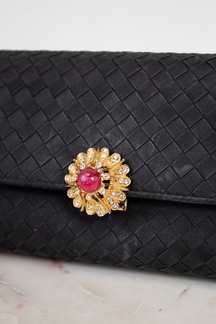 Black woven clutch with a decorative brooch featuring a pink gemstone and gold accents on a light background.