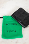 Black woven wallet with Bottega Veneta logo and green drawstring pouch on a light marble background