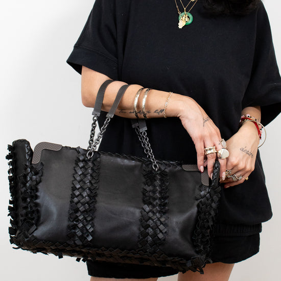 Person holding a black handbag with braided details against a plain background
