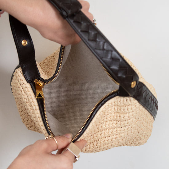 Woven handbag with black leather trim held by a person against a neutral background