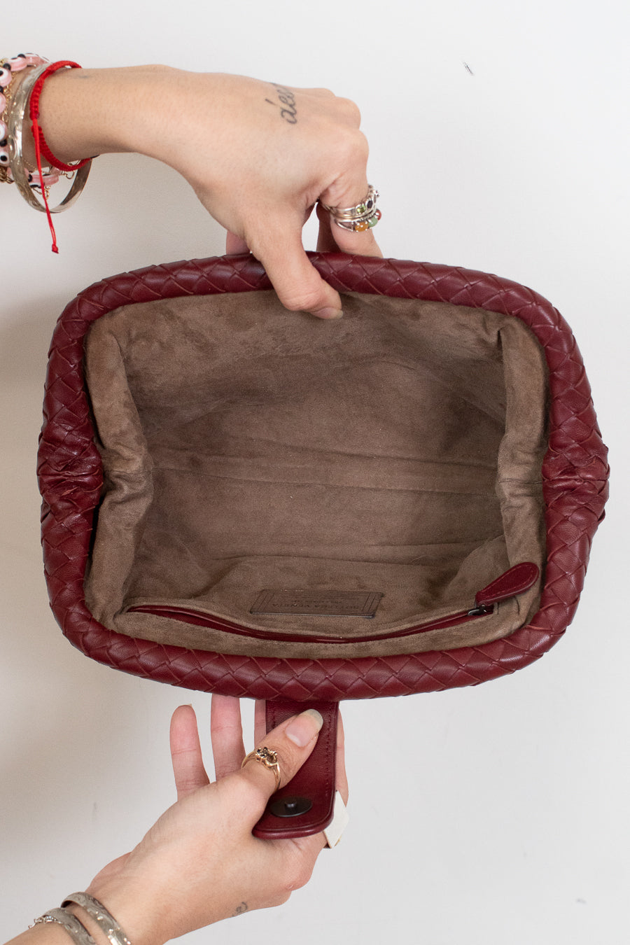 A close-up of a burgundy quilted leather handbag held open by two hands against a plain white background, showing a brown suede-like interior lining.