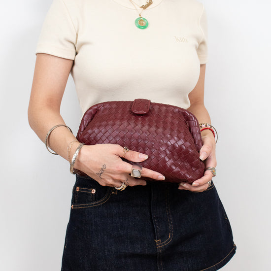 A woman wearing a cream short-sleeve top and a dark denim mini skirt holds a burgundy woven leather clutch bag against a plain white background.