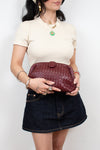A woman wearing a cream short-sleeve top and a dark denim mini skirt holds a burgundy woven leather clutch bag against a plain white background.