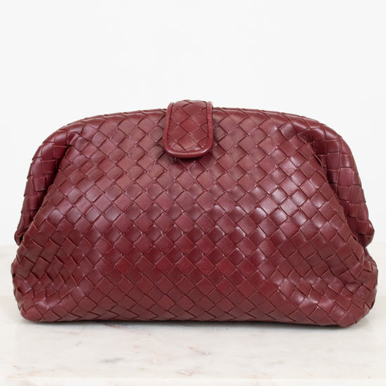 A woven leather clutch in a deep maroon, featuring a top snap closure, displayed on a white marble surface against a white background.