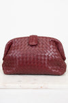 A woven leather clutch in a deep maroon, featuring a top snap closure, displayed on a white marble surface against a white background.