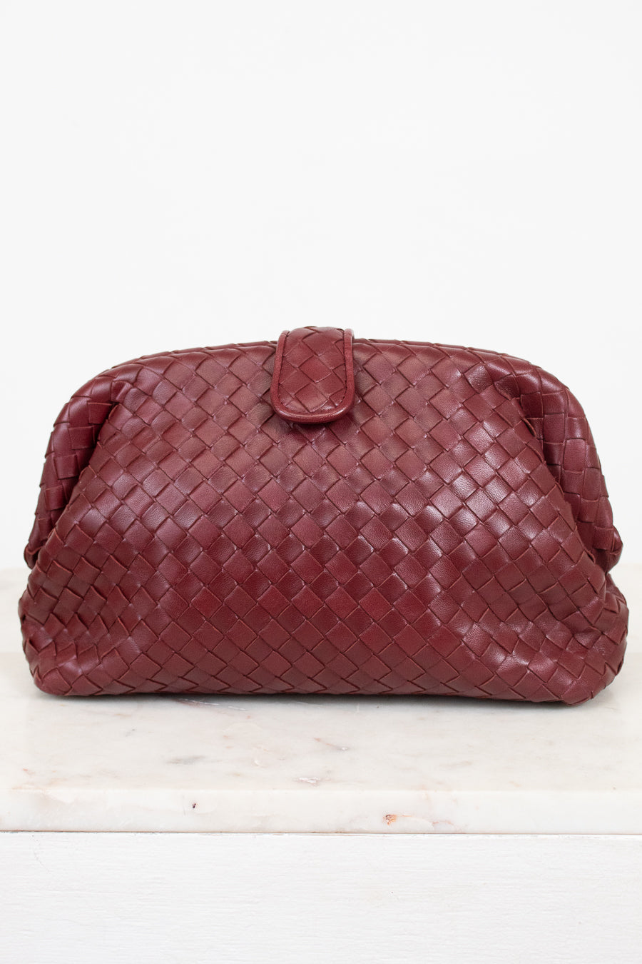 A woven leather clutch in a deep maroon, featuring a top snap closure, displayed on a white marble surface against a white background.