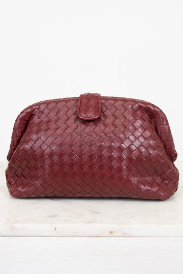 A woven leather clutch in a deep maroon, featuring a top snap closure, displayed on a white marble surface against a white background.