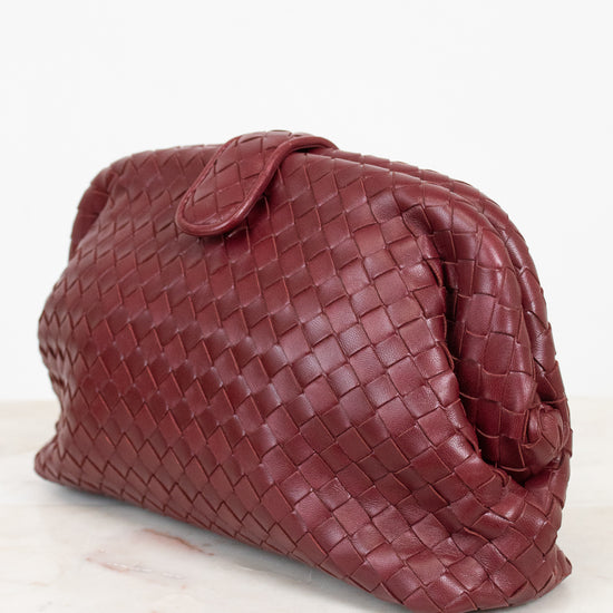 A dark burgundy woven leather clutch bag with a top snap closure, resting on a white marble surface against a white background.