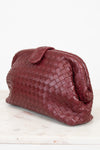 A dark burgundy woven leather clutch bag with a top snap closure, resting on a white marble surface against a white background.