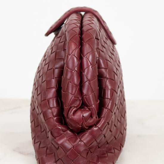A close-up of a woven leather handbag in a deep burgundy red, showing the detailed basketweave texture and top snap closure against a white background.