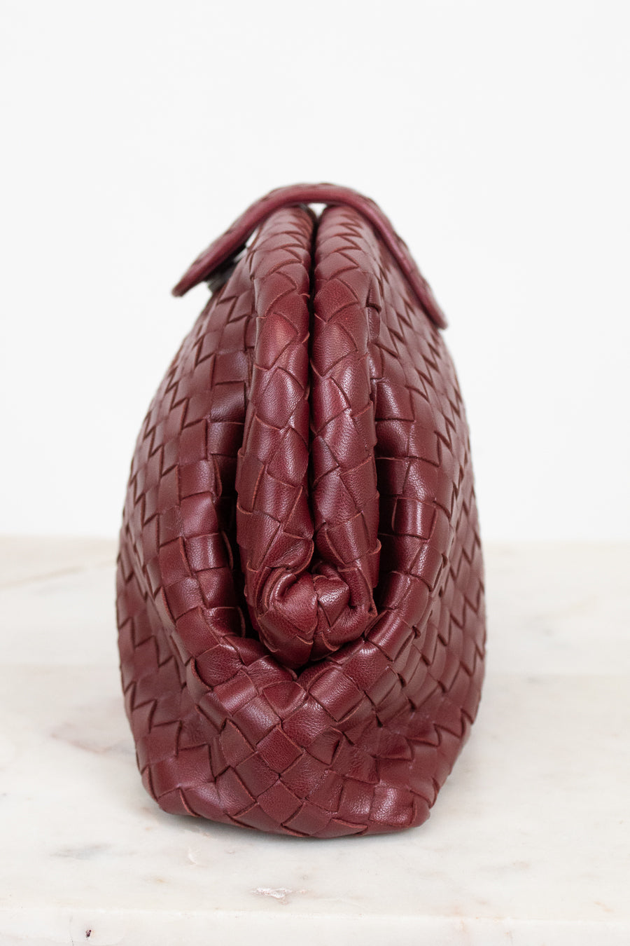A close-up of a woven leather handbag in a deep burgundy red, showing the detailed basketweave texture and top snap closure against a white background.