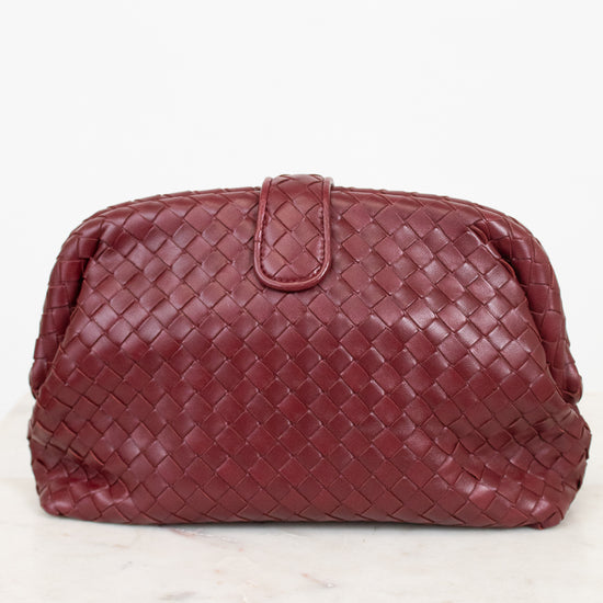 A woven leather clutch in a deep burgundy red, featuring a top snap closure, displayed on a white marble surface against a white background.