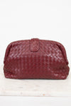 A woven leather clutch in a deep burgundy red, featuring a top snap closure, displayed on a white marble surface against a white background.