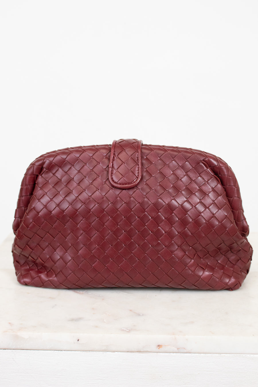 A woven leather clutch in a deep burgundy red, featuring a top snap closure, displayed on a white marble surface against a white background.
