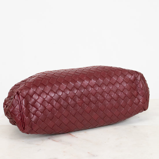 A woven leather clutch in a deep burgundy red, resting on a white marble surface against a white background.