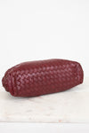 A woven leather clutch in a deep burgundy red, resting on a white marble surface against a white background.