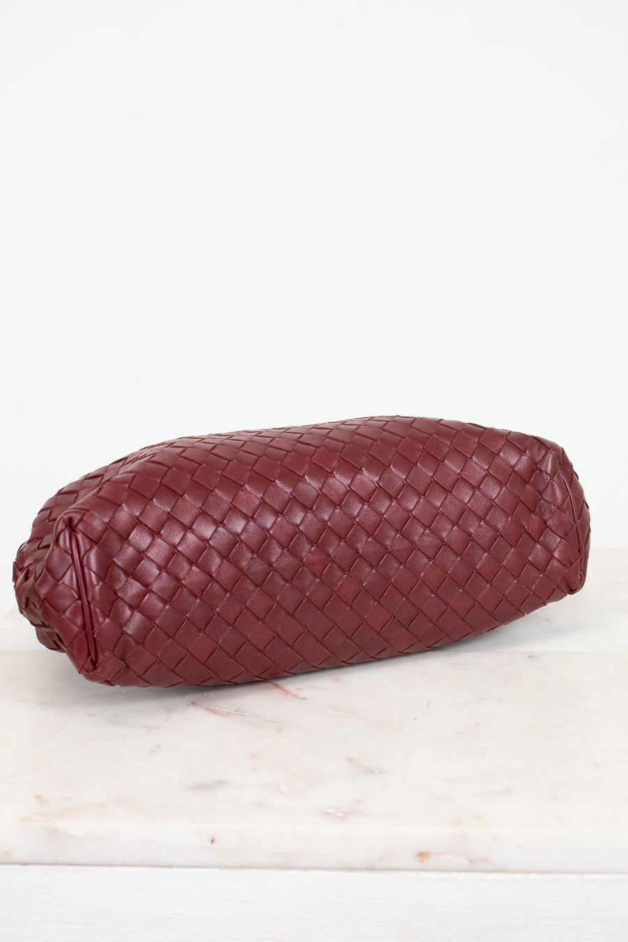A woven leather clutch in a deep burgundy red, resting on a white marble surface against a white background.