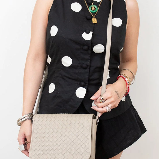 Woman wearing a black dress with white polka dots and holding a beige handbag against a plain background