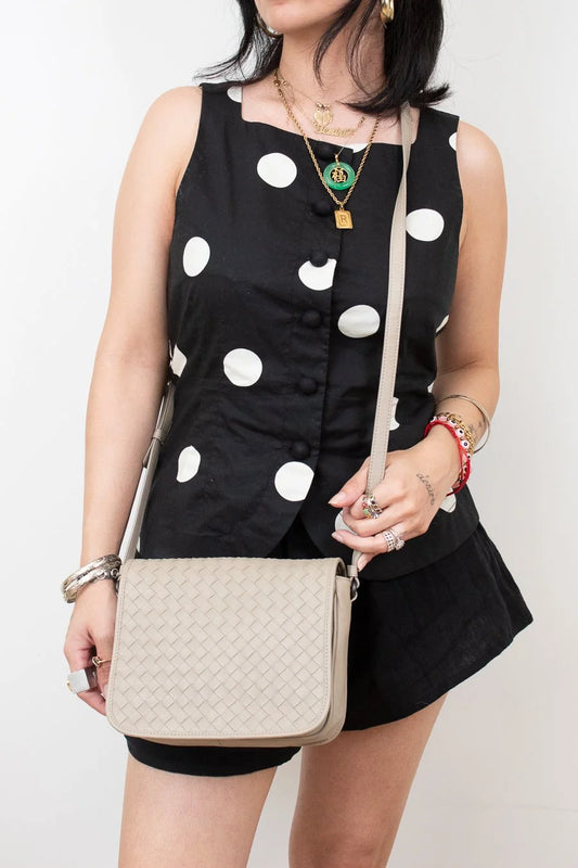 Woman wearing a black dress with white polka dots and holding a beige handbag against a plain background