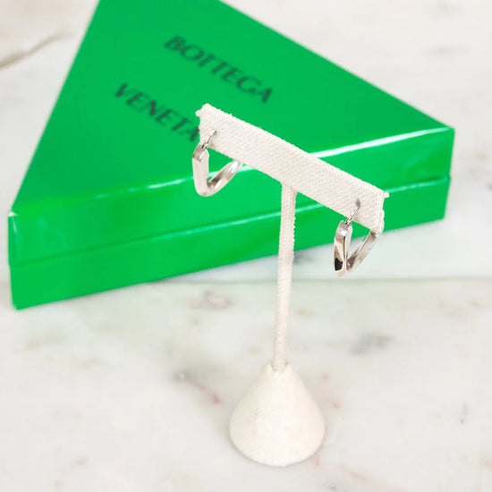 Silver earring stand with two silver earrings in front of a green Bottega Veneta box on a marble surface.