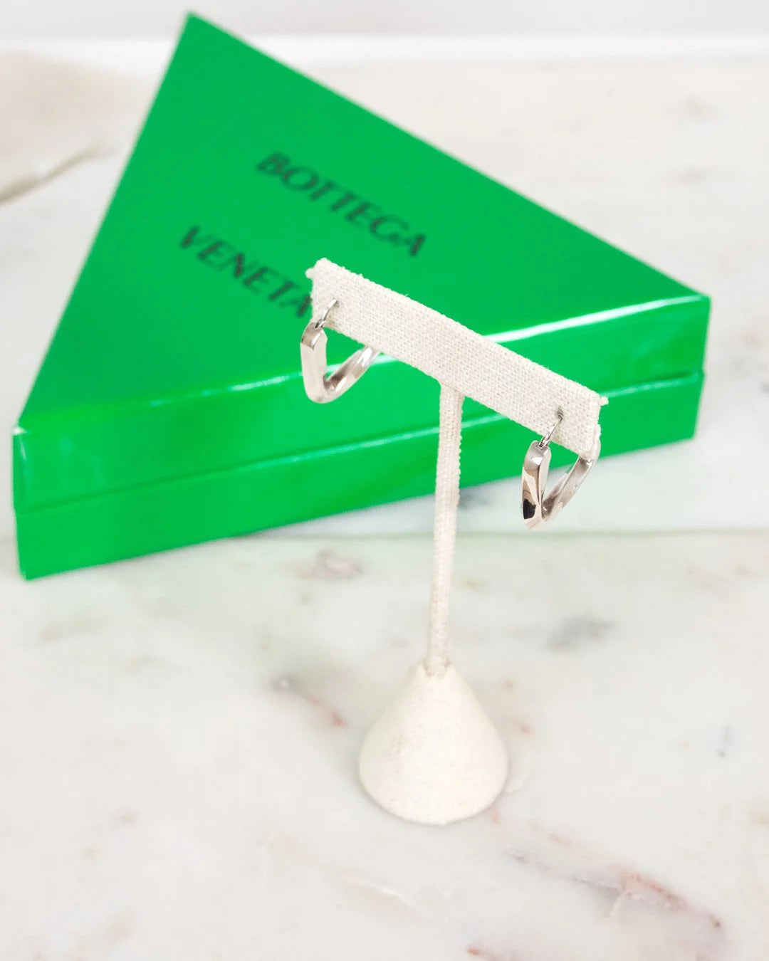 Silver earring stand with two silver earrings in front of a green Bottega Veneta box on a marble surface.