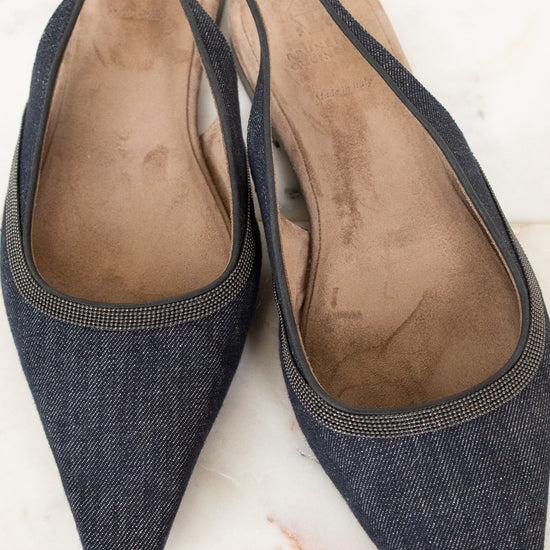A pair of pointed-toe slingback flats in dark blue denim fabric with a thin metallic trim, shown on a light marble surface.