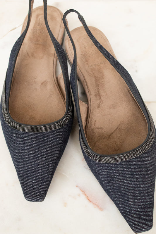 A pair of pointed-toe slingback flats in dark blue denim fabric with a thin metallic trim, shown on a light marble surface.