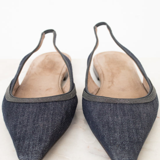 A pair of pointed-toe slingback flats made from dark blue denim, featuring a thin metallic trim along the edge and a light tan insole, photographed on a light marble surface.