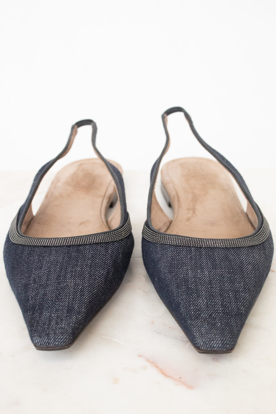 A pair of pointed-toe slingback flats made from dark blue denim, featuring a thin metallic trim along the edge and a light tan insole, photographed on a light marble surface.