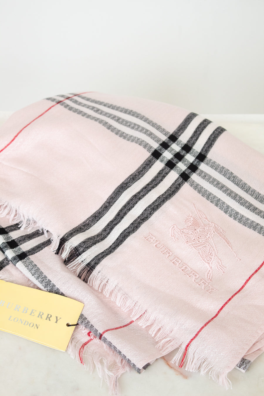 Pink plaid scarf with black and red stripes on a white background, featuring the Burberry logo.