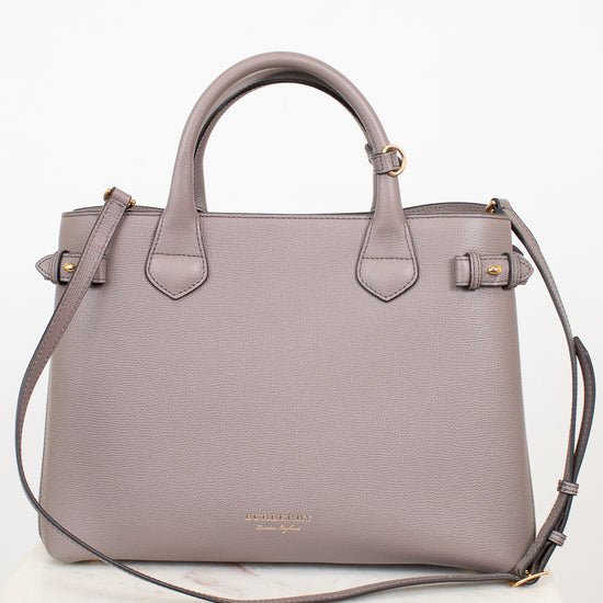 A structured taupe leather handbag with double top handles, a shoulder strap, and gold-tone hardware, displayed on a white marble surface against a white background.