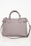 A structured taupe leather handbag with double top handles, a shoulder strap, and gold-tone hardware, displayed on a white marble surface against a white background.