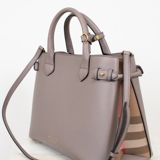 A taupe leather handbag with gold-tone hardware and a signature check-patterned side panel, displayed on a light marble surface against a white background.