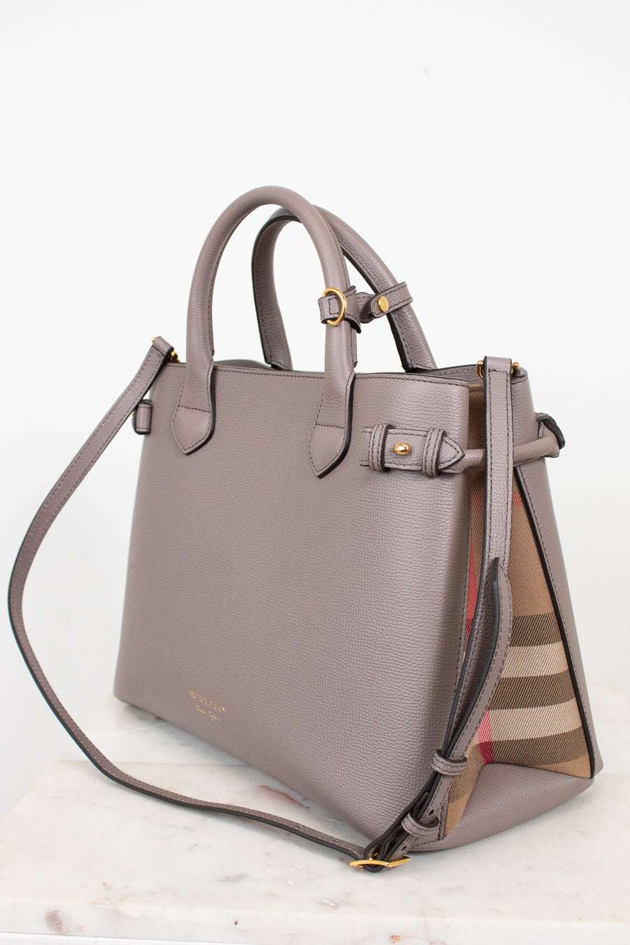 A taupe leather handbag with gold-tone hardware and a signature check-patterned side panel, displayed on a light marble surface against a white background.