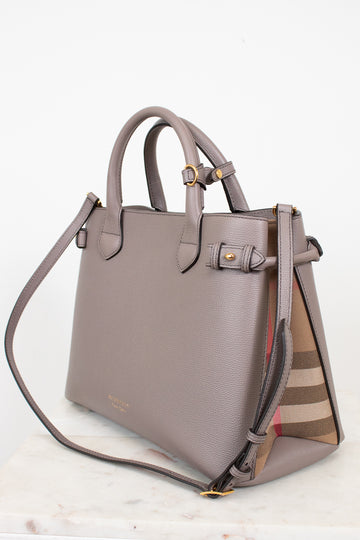 A taupe leather handbag with gold-tone hardware and a signature check-patterned side panel, displayed on a light marble surface against a white background.