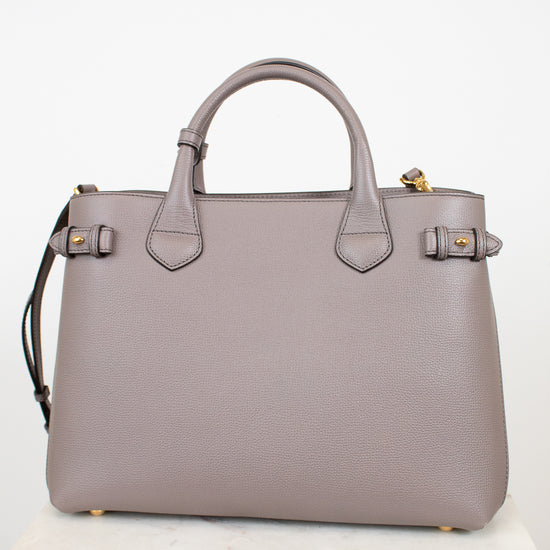A structured taupe leather handbag with double top handles, gold-tone hardware, and a shoulder strap, displayed on a light marble surface against a white background.