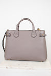 A structured taupe leather handbag with double top handles, gold-tone hardware, and a shoulder strap, displayed on a light marble surface against a white background.