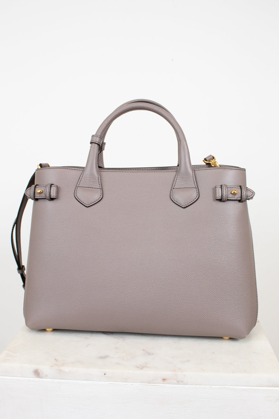 A structured taupe leather handbag with double top handles, gold-tone hardware, and a shoulder strap, displayed on a light marble surface against a white background.