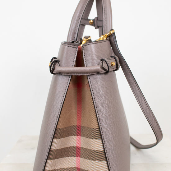 A structured leather handbag in taupe with a signature beige, brown, and red check pattern panel on the front, gold-tone hardware, and a shoulder strap, photographed against a clean white background.