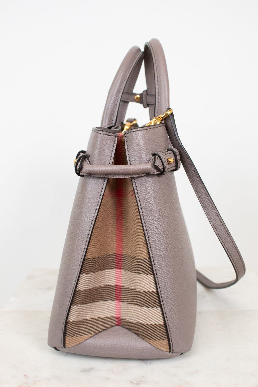 A structured leather handbag in taupe with a signature beige, brown, and red check pattern panel on the front, gold-tone hardware, and a shoulder strap, photographed against a clean white background.
