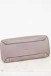 The bottom base of a taupe pebbled leather handbag featuring gold-tone metal studs on the base, resting on a white marble surface against a white background.