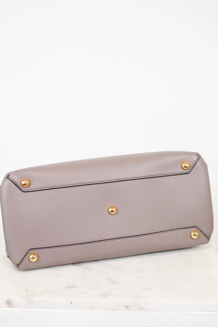 The bottom base of a taupe pebbled leather handbag featuring gold-tone metal studs on the base, resting on a white marble surface against a white background.