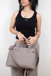 A woman wearing a black ribbed tank top and light gray sweatpants holds a large taupe leather shoulder bag with gold-tone hardware and a gold Burberry logo. 