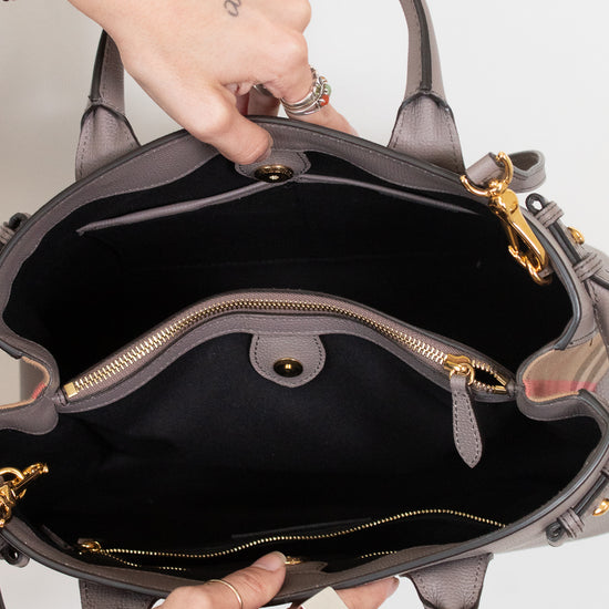 An open taupe leather handbag with gold-tone hardware, shown from the inside to display its black fabric lining, zippered compartments, and interior layout. A person's hands with tattoos are holding the bag open against a plain white background.