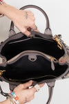 An open taupe leather handbag with gold-tone hardware, shown from the inside to display its black fabric lining, zippered compartments, and interior layout. A person's hands with tattoos are holding the bag open against a plain white background.