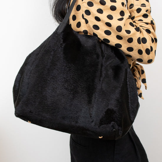 A person wearing a tan and black polka dot blouse carries a large black fuzzy shoulder bag against a plain light background.