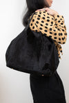 A person wearing a tan and black polka dot blouse carries a large black fuzzy shoulder bag against a plain light background.